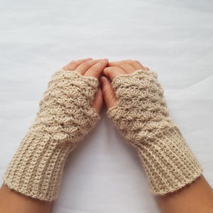 May include: A pair of beige fingerless gloves, crocheted with a textured pattern. The gloves have ribbed cuffs and a shell-like design on the hand portion. The neutral color makes them versatile for various outfits.