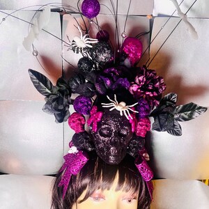 May include: A Halloween headpiece with a black glitter skull, purple and black floral details, and decorative spiders. The headpiece includes purple glitter bows and decorative accents, ideal for a costume or themed event.