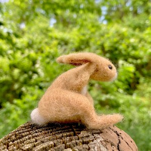 May include: A small, brown, felt bunny rabbit sitting on a tree stump. The bunny has a white tail and is looking to the right.
