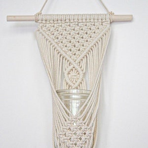 May include: A macrame plant hanger with a wooden dowel and a glass jar. The hanger is made of natural cotton cord and has a triangular shape with a fringe at the bottom.