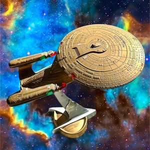 May include: A gold-colored model of the Starship Enterprise NCC-1701-E featuring a large circular saucer section, two elongated warp nacelles with red and green accents, and a metallic display stand with the Star Trek logo on a circular base.
