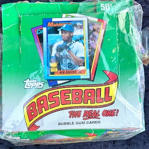 May include: Sealed box of Topps Baseball cards. The box is green with the words "BASEBALL THE REAL ONE! BUBBLE GUM CARDS" in red and yellow. A baseball card featuring Ken Griffey Jr. is visible. The box is wrapped in clear plastic.