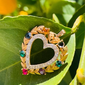 May include: A gold-toned heart-shaped pendant with a clear rhinestone border. The heart is surrounded by a wreath of gold leaves and colorful gemstones, including blue, green, red, and orange. The pendant is set against a green leaf background.