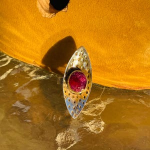 Puede incluir: Anillo de plata y oro con diseño ovalado. El anillo presenta una gema roja vibrante engastada en un bisel dorado. La parte plateada tiene un patrón texturizado de pequeños agujeros espaciados uniformemente.
