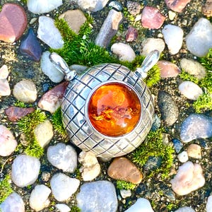 May include: A round silver pendant featuring a textured grid pattern across its surface and a large, polished, translucent orange amber cabochon set in the center. The pendant includes two silver loops at the top for attachment to a chain.