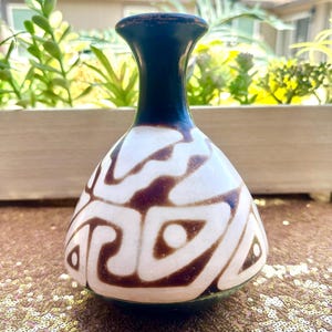 Puede incluir: Un pequeño jarrón decorativo con cuello azul oscuro y cuerpo blanco adornado con patrones abstractos marrones. El jarrón tiene una base redondeada y una abertura estrecha, adecuado para exhibir flores pequeñas o como adorno decorativo.