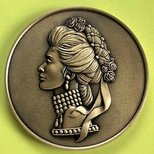 May include: A bronze-colored round medallion featuring a detailed profile of a woman with an elaborate hairstyle adorned with roses. The woman wears a necklace and earrings. The medallion has a textured background.