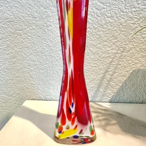 Puede incluir: Un jarrón alto de cristal rojo con base acampanada y cintura estrecha. El jarrón está decorado con salpicaduras abstractas y coloridas de amarillo, blanco, azul y verde. El jarrón mide unos 25 cm de alto.
