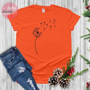 May include: Orange t-shirt with a black dandelion graphic. The dandelion is blowing in the wind and has seeds floating away.