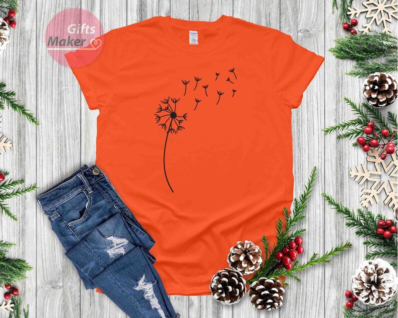 May include: Orange t-shirt with a black dandelion graphic. The dandelion is blowing in the wind and has seeds floating away.
