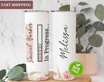 May include: Two white stainless steel tumblers with a floral design. One tumbler says "Social Worker... In Progress" with a progress bar. The other tumbler says "Melissa".