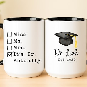 May include: White ceramic mug with a black handle and rim. One side features a checklist with "It's Dr. Actually" checked. The other side displays a graduation cap graphic, the name "Dr. Leah", and "Est. 2025".