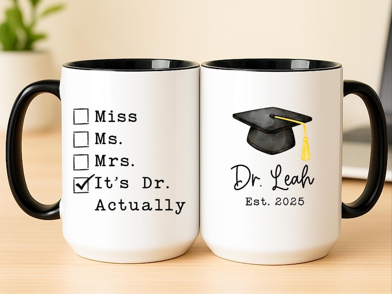 May include: White ceramic mug with a black handle and rim. One side features a checklist with "It's Dr. Actually" checked. The other side displays a graduation cap graphic, the name "Dr. Leah", and "Est. 2025".