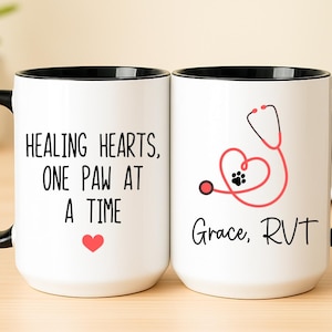 May include: Two white ceramic mugs with black handles and rims. One mug has the text "HEALING HEARTS, ONE PAW AT A TIME" with a red heart. The other mug features a stethoscope forming a heart with a paw print and the text "Grace, RVT".