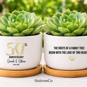 May include: Two white ceramic planters with bamboo saucers, each containing a green succulent. One planter features gold text reading 50th ANNIVERSARY, Sarah & Ethan, and 08.12.1976. The other displays THE ROOTS OF A FAMILY TREE BEGIN WITH THE LOVE OF TWO HEARTS.