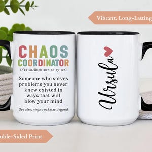 May include: White ceramic mug with a black handle and rim. The mug features a colourful graphic on one side that reads "Chaos Coordinator" with a definition and the text "See also: ninja, rockstar, legend". The other side of the mug has the name "Ursula" in black cursive with a small pink heart above it. The mug is on a white surface.