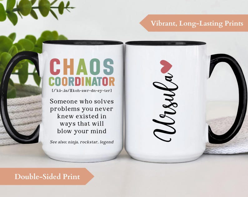 May include: White ceramic mug with a black handle and rim. The mug features a colourful graphic on one side that reads "Chaos Coordinator" with a definition and the text "See also: ninja, rockstar, legend". The other side of the mug has the name "Ursula" in black cursive with a small pink heart above it. The mug is on a white surface.
