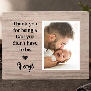 May include: A light-colored wooden picture frame with a photograph of a father kissing a baby. The frame has the text "Thank you for being a Dad you didn't have to be." and the name "Sheryl".