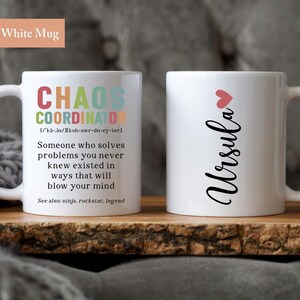 May include: Two white mugs, one with the text "CHAOS COORDINATOR" and a definition, and the other with the name "Ursula" in cursive with a small pink heart above it.