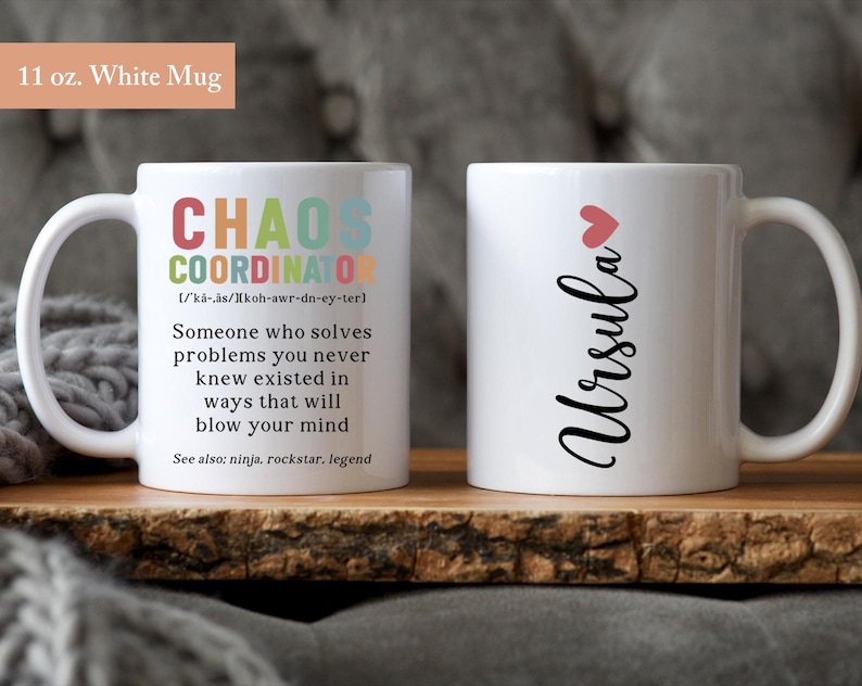 May include: Two white mugs, one with the text "CHAOS COORDINATOR" and a definition, and the other with the name "Ursula" in cursive with a small pink heart above it.