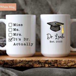 May include: Two white ceramic mugs. One mug has the text "Miss, Ms., Mrs., It's Dr. Actually". The other mug has a graduation cap graphic, the name "Dr. Leah", and "Est. 2025". Each mug is 15 oz.
