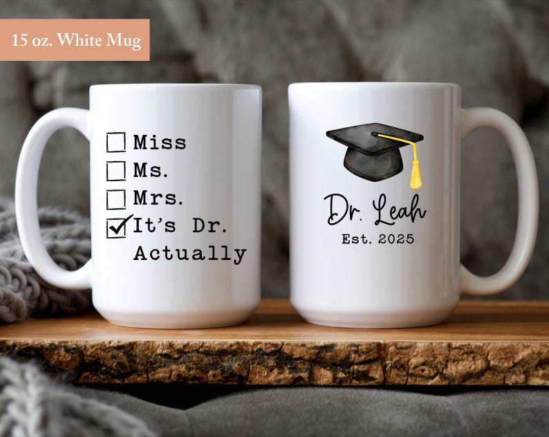May include: Two white ceramic mugs. One mug has the text "Miss, Ms., Mrs., It's Dr. Actually". The other mug has a graduation cap graphic, the name "Dr. Leah", and "Est. 2025". Each mug is 15 oz.