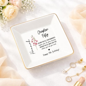 May include: A white ceramic trinket dish with gold trim, featuring the text "Chapter Fifty" and a floral design. The dish includes the name "Hannah" and a birthday message. A decorative and personalized gift.