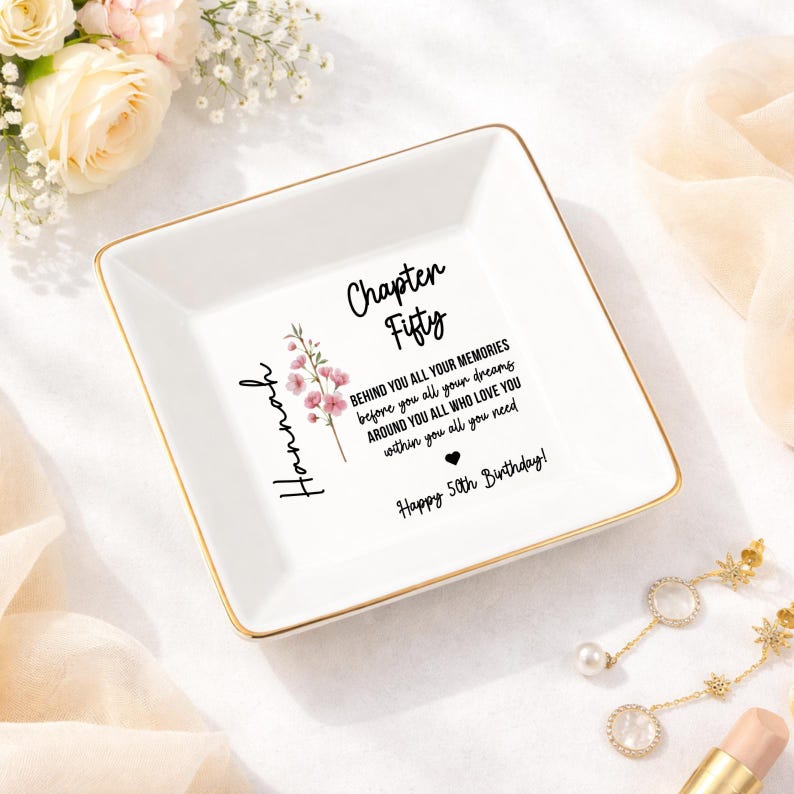 May include: A white ceramic trinket dish with gold trim, featuring the text "Chapter Fifty" and a floral design. The dish includes the name "Hannah" and a birthday message. A decorative and personalized gift.