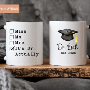 May include: Two white ceramic mugs with black handles and rims. One mug has a checklist with "It's Dr. Actually" checked. The other mug features a graduation cap graphic and the name "Dr. Leah Est. 2025".