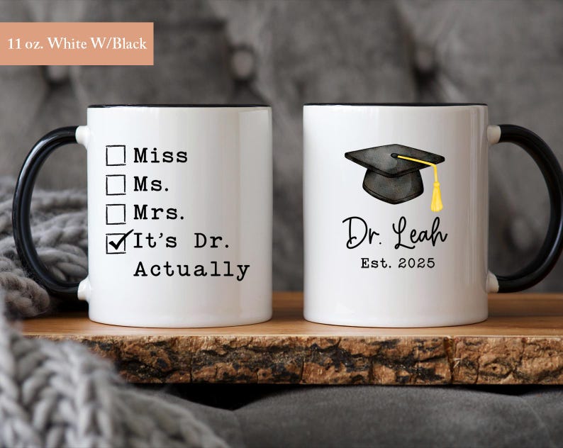 May include: Two white ceramic mugs with black handles and rims. One mug has a checklist with "It's Dr. Actually" checked. The other mug features a graduation cap graphic and the name "Dr. Leah Est. 2025".