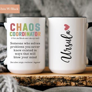 May include: White ceramic mug with a black handle and rim. The mug features a black and white design with the text "Chaos Coordinator" and a definition. The other side of the mug has the name "Ursula" in black cursive with a pink heart.