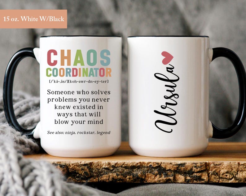 May include: White ceramic mug with a black handle and rim. The mug features a black and white design with the text "Chaos Coordinator" and a definition. The other side of the mug has the name "Ursula" in black cursive with a pink heart.