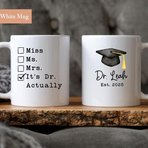 May include: Two white ceramic mugs. One mug has the text "Miss, Ms., Mrs., It's Dr. Actually". The other mug has a graduation cap graphic, the name "Dr. Leah", and "Est. 2025". Each mug is 11 oz.