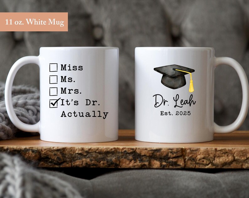 May include: Two white ceramic mugs. One mug has the text "Miss, Ms., Mrs., It's Dr. Actually". The other mug has a graduation cap graphic, the name "Dr. Leah", and "Est. 2025". Each mug is 11 oz.