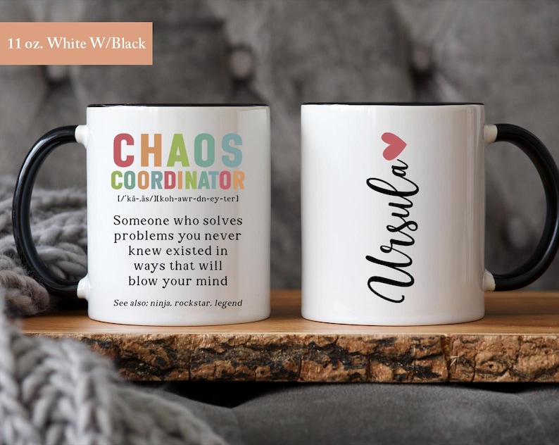 May include: Two 11 ounce white ceramic mugs with black handles and black rims. One mug has the text "CHAOS COORDINATOR" with a definition and the text "See also: ninja, rockstar, legend". The other mug has the text "Ursula" with a pink heart above it.