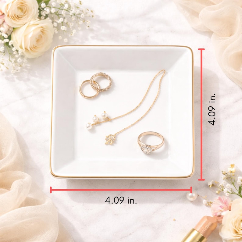 May include: A white square jewelry tray with gold trim, measuring 4.09 inches on each side. The tray holds gold rings and a necklace with pearl accents. The background includes flowers and a soft, neutral fabric.