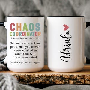 May include: Two white ceramic mugs with black handles. One mug has the text "CHAOS COORDINATOR" with a definition and the text "See also: ninja, rockstar, legend". The other mug has the text "Ursula" with a pink heart above it.