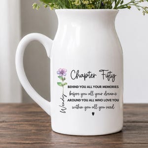 May include: A white ceramic pitcher mug featuring a floral illustration of a purple flower and the text: Chapter Fifty, BEHIND YOU ALL YOUR MEMORIES, before you all your dreams, AROUND YOU ALL WHO LOVE YOU, within you all you need, and the name Wendy.