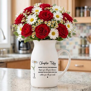 May include: A white ceramic pitcher vase filled with a bouquet of red roses and white daisies. The pitcher features black printed text that reads Chapter Fifty, followed by sentimental phrases and the name Sarah, accompanied by a daisy illustration.
