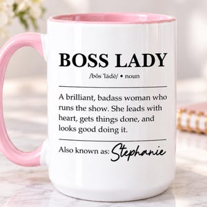 May include: A white ceramic mug with a light pink handle and interior rim. The front features black text reading BOSS LADY, a phonetic pronunciation, and a definition describing a leader. The bottom includes the text Also known as: Stephanie.