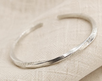 Puede incluir: Una pulsera abierta de tono plateado con una superficie texturizada y martillada, con una forma ligeramente retorcida e irregular. El metal tiene un acabado reflectante que capta la luz, resaltando los detalles orgánicos a lo largo de la banda.