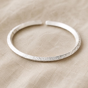 Puede incluir: Una pulsera abierta de color plateado con una superficie exterior texturizada y martillada, y extremos lisos y redondeados. La banda circular tiene un acabado met&aacute;lico reflectante y un ancho constante en todo su dise&ntilde;o.