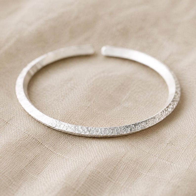 Puede incluir: Una pulsera abierta de color plateado con una superficie exterior texturizada y martillada, y extremos lisos y redondeados. La banda circular tiene un acabado met&aacute;lico reflectante y un ancho constante en todo su dise&ntilde;o.