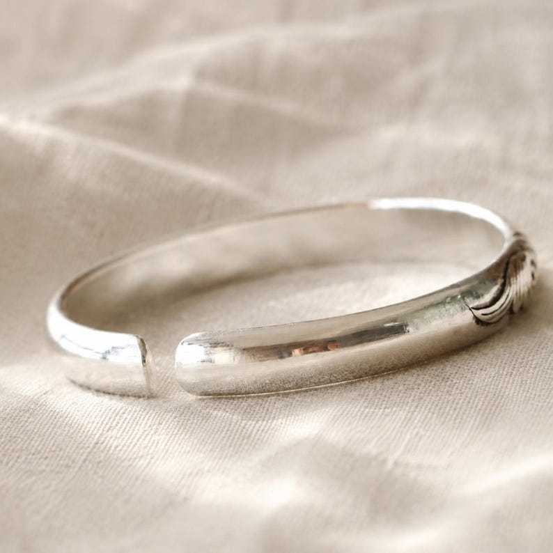 Puede incluir: Una pulsera de brazalete de plata pulida con una banda lisa y redondeada y un peque&ntilde;o detalle grabado en forma de ala en un lado. La pulsera presenta un dise&ntilde;o abierto que permite el ajuste y tiene un acabado met&aacute;lico reflectante.