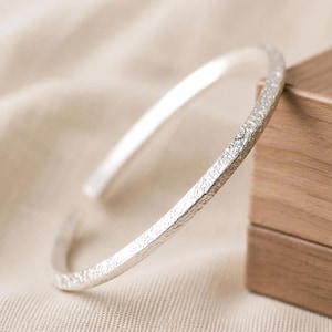 Puede incluir: Una pulsera abierta de color plateado con una superficie texturizada e irregular. El brazalete tiene un perfil circular delgado con una abertura en un extremo, mostrando un acabado met&aacute;lico que refleja la luz sobre su textura exterior granulada.