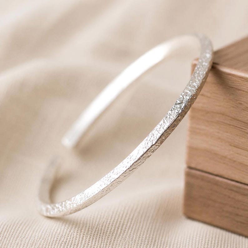 Puede incluir: Una pulsera abierta de color plateado con una superficie texturizada e irregular. El brazalete tiene un perfil circular delgado con una abertura en un extremo, mostrando un acabado met&aacute;lico que refleja la luz sobre su textura exterior granulada.