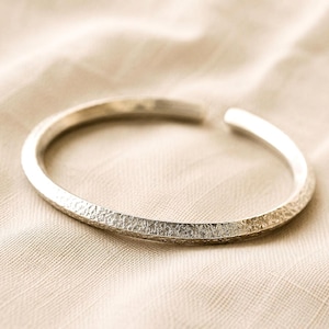 Puede incluir: Una pulsera abierta de color plateado con una superficie exterior texturizada y martillada. La banda de metal tiene un perfil redondeado y una abertura en un lado, mostrando un acabado met&aacute;lico que refleja la luz sobre su forma curva.