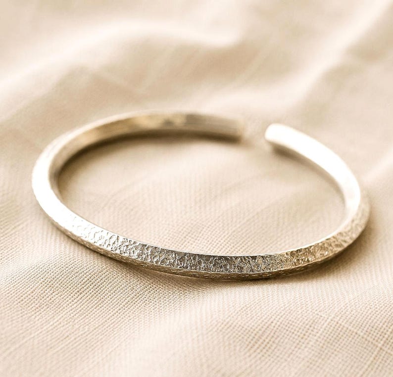Puede incluir: Una pulsera abierta de color plateado con una superficie exterior texturizada y martillada. La banda de metal tiene un perfil redondeado y una abertura en un lado, mostrando un acabado met&aacute;lico que refleja la luz sobre su forma curva.