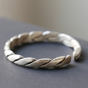 Può includere: Un braccialetto in argento con un design intrecciato. Il braccialetto è aperto e ha uno spazio su un lato.