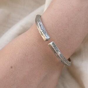 May include: A silver-toned open cuff bracelet featuring an oxidized finish and engraved floral patterns along the outer surface. The metal band has a rectangular profile with rounded edges and a gap at the center, showcasing intricate line work.